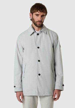 Man wearing a light gray buttoned coat with side pockets and buttoned cuffs, standing against a plain white background.