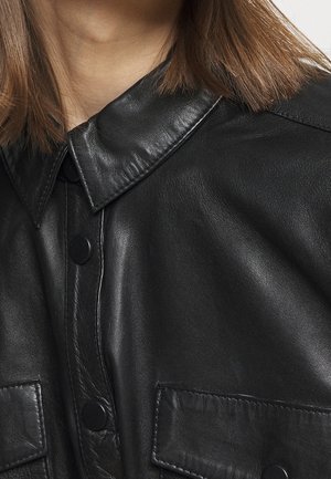 Femme portant une chemise boutonnée en cuir noir avec des poches à rabat et des cheveux bruns raides reposant sur les épaules.