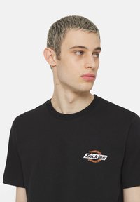 Camiseta de algodón negra con cuello redondo, que presenta un logo en blanco y naranja de "Dickies" en el lado izquierdo del pecho. Mangas cortas, corte estándar.
