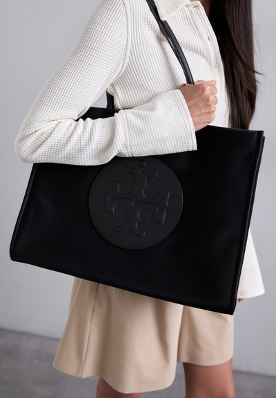 Sort tote-bag lavet af glat materiale, med et cirkulært præget logo, og to skulderremme, holdt af en person iført en hvid struktureret top.