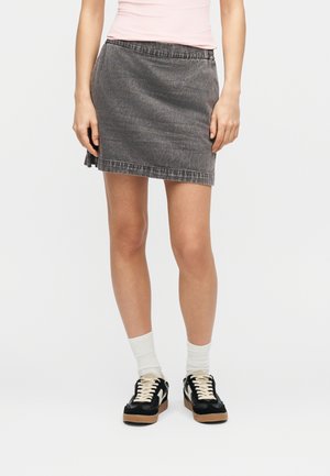 Person iført en falmet sort denim-mininederdel, hvide sokker, sorte sneakers med hvide snørebånd og en lyserød top, stående på en hvid baggrund.