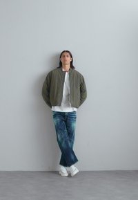 Olive rohelise bomber-jakk, valge T-särk, sidrunierror sinised teksad ja valged tossud. Modell toetub hallile seinale minimalistlikus keskkonnas.