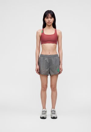 Ung kvinde stående med fronten mod kameraet iført en rød sports-bh, grå træningsshorts og hvide løbesko på en ensfarvet baggrund.