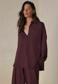 Burgundy button-up shirt with a pocket, smooth texture, and relaxed fit, paired with matching burgundy pants and a lace camisole underneath.