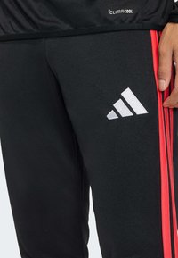 Μαύρο αθλητικό παντελόνι με κόκκινες πλευρικές ρίγες, λευκό λογότυπο Adidas και διαπνέον ύφασμα ClimaCool για διαχείριση της υγρασίας.