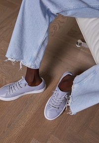 Gambe che indossano jeans azzurri sfilacciati e sneakers viola chiaro, sedute su una sedia bianca con sfondo in parquet.