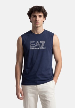 Uomo che indossa una maglietta smanicata blu navy con il logo "EA7 Emporio Armani", abbinata a pantaloni bianchi, in piedi su uno sfondo chiaro semplice.