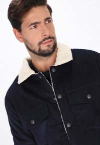 Marineblaues Cordjackett mit cremefarbenem Sherpa-Kragen, ausgestattet mit zwei Klapptaschen und metallischen Druckknöpfen. Strukturierter Stoff und lässiges Design.