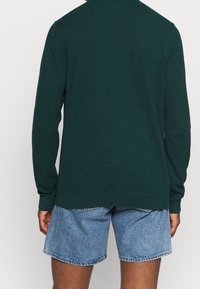 Mörkgrön långärmad tröja med rundad krage, ribbade ärmslut och nederkant. Bärs med ljusblå denimshorts. Strukturerad, avslappnad design.