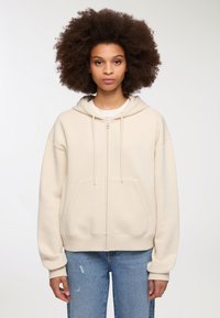 Jasno-beżowa bluza z kapturem zapinana na suwak, wykonana z miękkiego materiału, z kieszenią kangurzą, sznurkami oraz ściągaczami. Noszona z niebieskimi dżinsami.