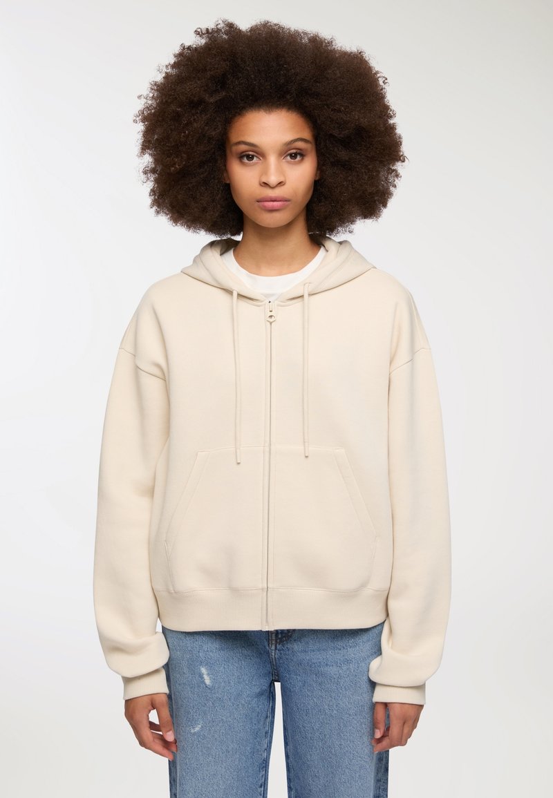Jasno-beżowa bluza z kapturem zapinana na suwak, wykonana z miękkiego materiału, z kieszenią kangurzą, sznurkami oraz ściągaczami. Noszona z niebieskimi dżinsami.