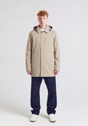 Junger Mann mit lockigem roten Haar, der einen beigen Kapuzenmantel, dunkelblaue Hosen und weiße Turnschuhe trägt, steht vor einem schlichten hellen Hintergrund.