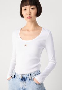 Haut blanc à manches longues côtelé avec un décolleté dégagé et un petit logo sur la poitrine, associé à un jean taille haute bleu clair.