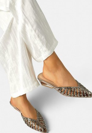 Voeten in puntige, open-toe platte mules met metalen studs, gecombineerd met witte, gestructureerde wijde broekspijpen.