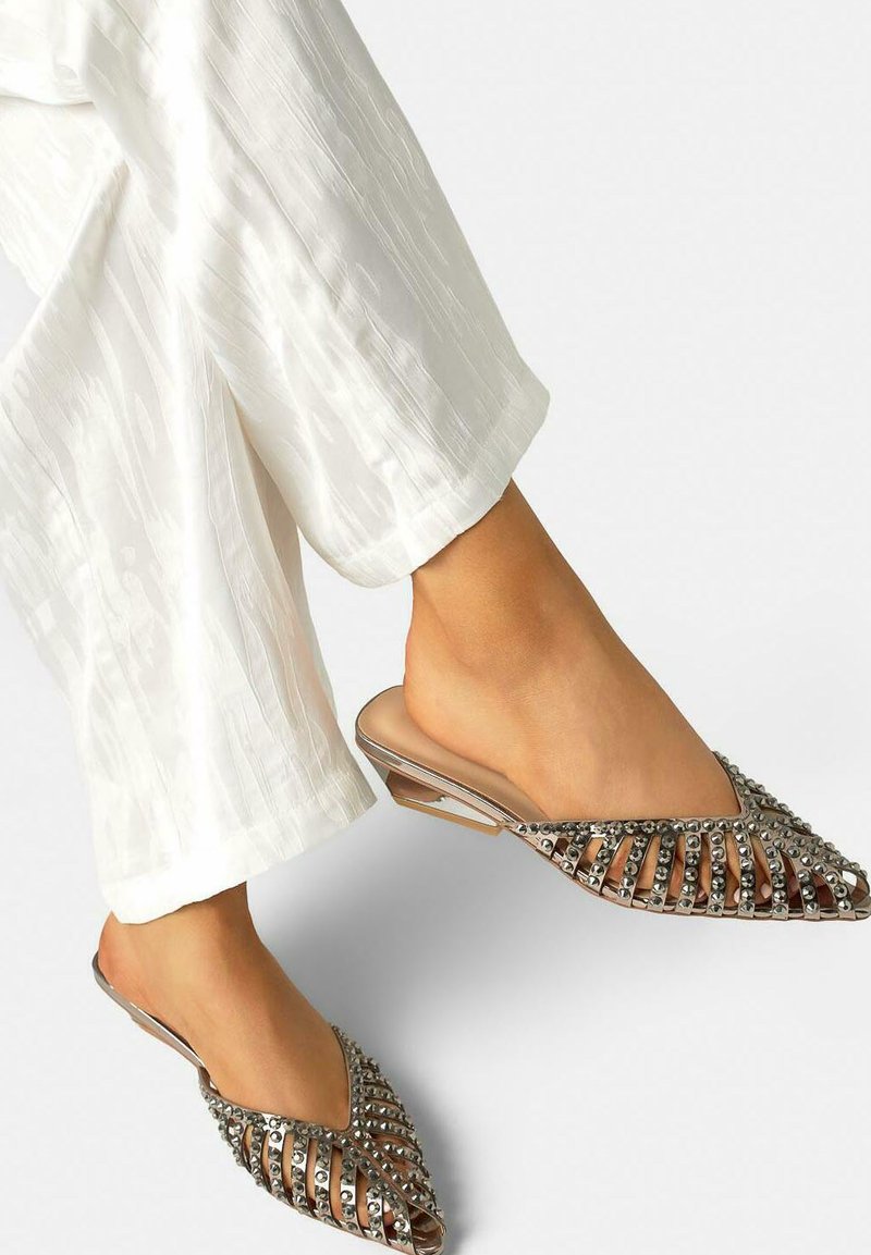 Pieds portant des mules plates ouvertes à bout pointu, ornées de clous métalliques, associées à un pantalon blanc à larges jambes texturé.