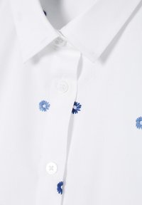 Witte polo t-shirt met blauwe geborduurde bloemaccenten. Beschikt over een klassieke kraag en een knopenfront. Zachte textuur met een subtiel weefpatroon.