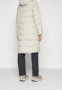 Lång, lättvärderad puffjacka i ljusbeige. Har horisontell sömnad, huva och en lätt lös passform. Bärs med svarta byxor och beige sneakers.