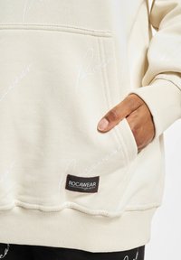 Krém színű kapucnis pulcsi fehér Rocawear logónyomattal, bordázott mandzsettákkal és elülső zsebből áll. Fekete Rocawear címke varrva az alján. Sima szövettextúra.