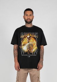 Sort bomulds T-shirt med et grafisk motiv af en skeletfigur mod en lys cirkulær baggrund, med lyn effekter og fed tekst.