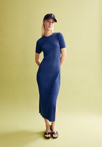 Ribbed, short-sleeve blue dress with side slit, paired with black sandals. Model wearing a black cap with a logo. Light green background.