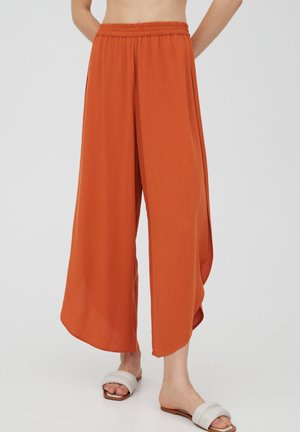 Personne portant un pantalon orange ample taille haute à jambes larges et des sandales blanches, debout devant un fond uni clair.