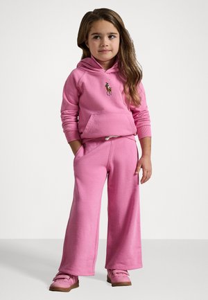 Rosa hettegenser med frontlomme og brodert logo, kombinert med vide rosa joggebukser. Antrekket har snøring og myk tekstur.