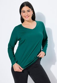 Dlouhý rukáv V-krk top v tmavě zelené barvě, vyrobený z měkké tkaniny, s uvolněným střihem a bočními rozparky, spárovaný s černými džíny.