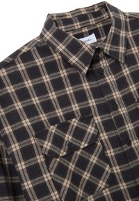 Camisa de franela a cuadros en gris oscuro y beige. Presenta un cuello con botones, bolsillo en el pecho y botones negros con una textura suave.