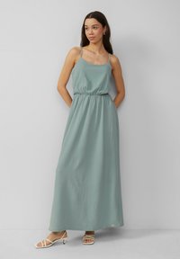 Maxikleid aus hellgrünem Stoff mit dünnen Trägern, einem elastischen Taillenbund und einem fließenden Design, kombiniert mit weißen Riemchensandalen.