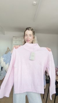 Maglione rosa lavorato a maglia con collo alto a coste e maniche lunghe. Presenta un motivo a rilievo e un'etichetta appesa al collo.