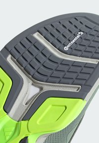 Semelle de chaussure présentant une texture en caoutchouc gris avec un motif de crantage surélevé, des accents jaune-vert et un détail de semelle intermédiaire en caoutchouc blanc.