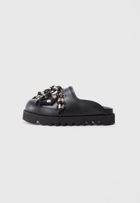 Black slip-on shoe featuring textured surface, decorative metal accents, and a thick, zigzag sole for added grip and style.