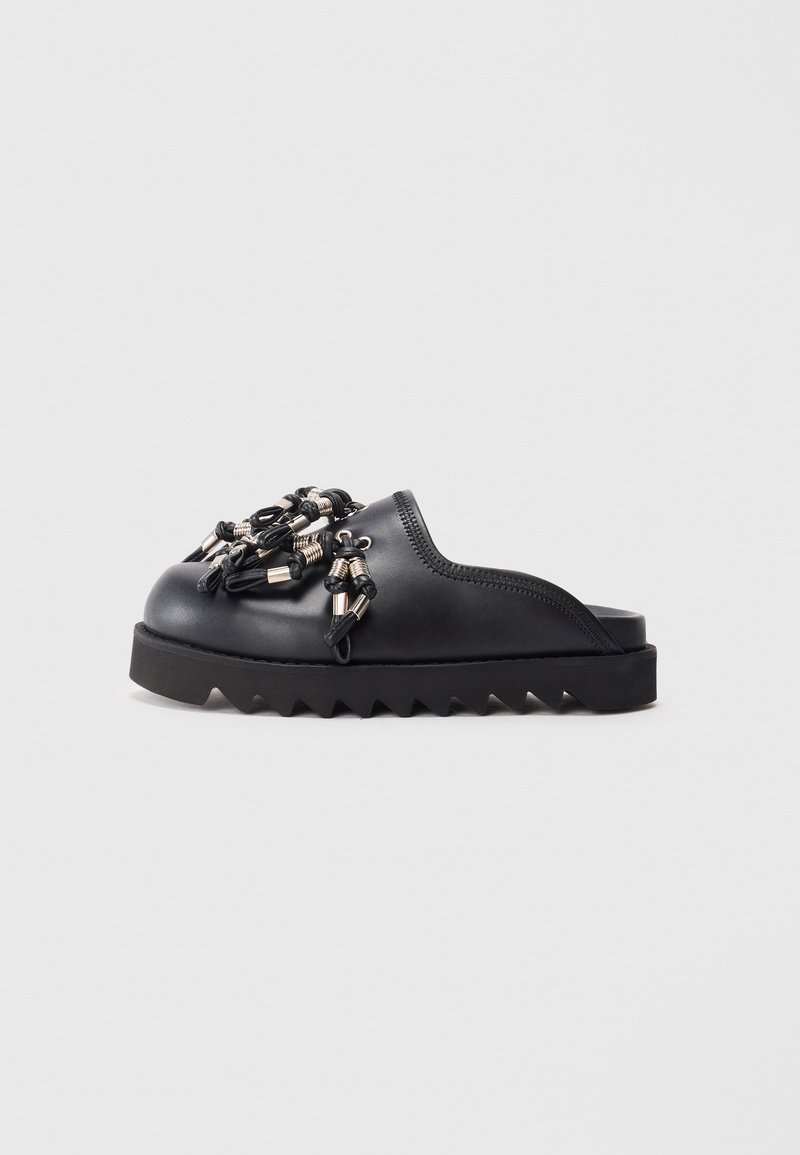 Black slip-on shoe featuring textured surface, decorative metal accents, and a thick, zigzag sole for added grip and style.