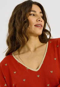 Top rojo de cuello en V con patrones de corazones dorados, con un ribete dorado brillante alrededor del escote. Textura suave y ajuste relajado.