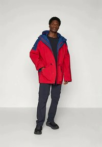 Chaqueta impermeable roja con capucha azul, que cuenta con bolsillos frontales, cordones ajustables y un corte relajado. Combinada con pantalones cargo oscuros.