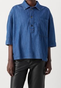 Denimhemd mit Kragen, kurzen Ärmeln, vorderem Knopfverschluss und einer Brusttasche. Blaue Farbe mit glatter Textur.