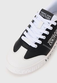 Svart og hvit sneaker med teksturert svart skinn og glatt hvitt materiale, med hvite snørebånd og "VERSACE JEANS COUTURE"-branding.