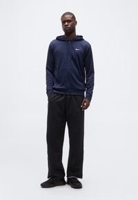 Νεαρός άνδρας στέκεται φορώντας μπλε σκούρο φούτερ με φερμουάρ Nike, μαύρο φαρδύ παντελόνι και μαύρα αθλητικά παπούτσια μπροστά σε απλό λευκό φόντο.