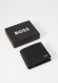 Melns bifolds maks, kas izgatavots no teksturēta materiāla, ar sudraba logotipu un tīrām līnijām, novietots blakus melnai kastei ar baltu logotipu.