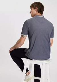 Szara koszulka polo z krótkim rękawem i białym wykończeniem przy kołnierzu i rękawach, noszona przez mężczyznę siedzącego na białym stołku, z tatuażem na ramieniu.