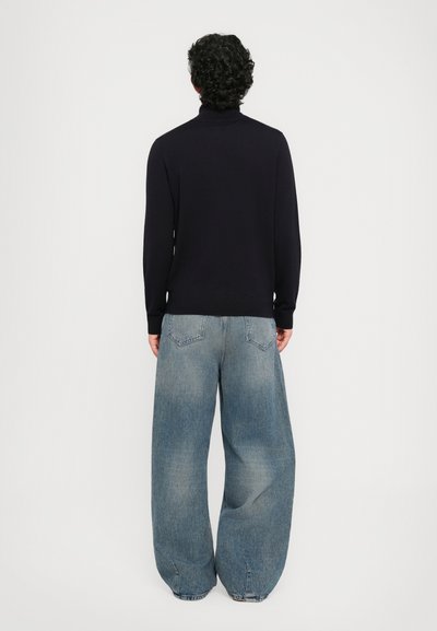 Person med lockigt hår iklädd en svart långärmad tröja och stora urblekta blå jeans, stående vänd bort mot en enfärgad bakgrund.