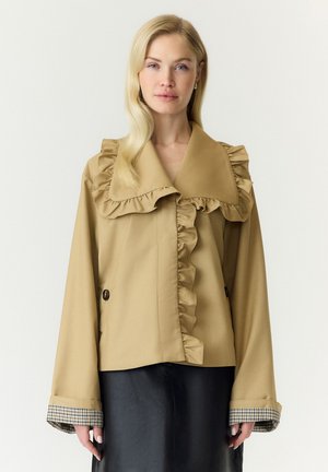Femme blonde porte une veste beige avec un grand col volanté et des poignets, associée à une jupe noire, debout devant un fond clair uni.