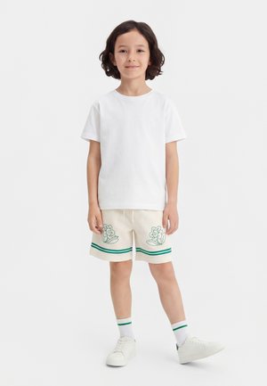 Παιδί όρθιο που φοράει απλό άσπρο μπλουζάκι, κρεμ σορτς με πράσινα σχέδια λουλουδιών, λευκές κάλτσες με πράσινες ρίγες και λευκά αθλητικά παπούτσια.