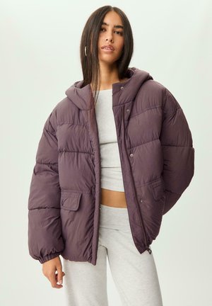 Žena oblečená v nadmernej purpurovej páperovej bunde s kapucňou, pod ktorou má na sebe svetlo šedý cropped top a tepláky, stojí pred jednoducho pozadím.