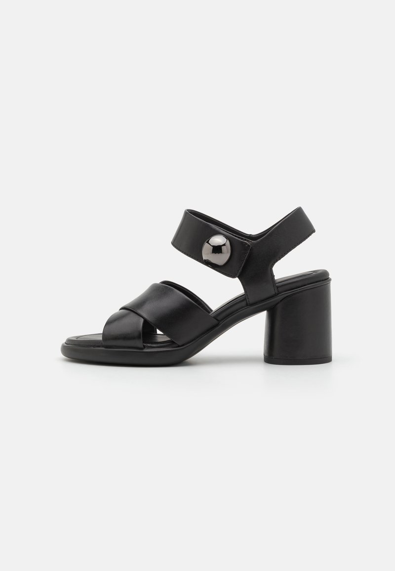 Sandale en cuir noir avec talon carré, bride de cheville ajustable ornée d'un grand bouton argenté, et lanières croisées à l'avant.