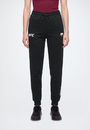 Modelo con pantalones de chándal negros de UFC, cintura con cordón, logotipos blancos, bajos ajustados en los tobillos y zapatillas deportivas negras.