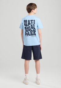 Camiseta de algodón azul claro con gran texto negro que dice "PARQUE NACIONAL", combinada con pantalones cortos azul marino. Zapatillas blancas y calcetines hasta la pantorrilla.