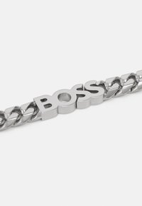 Hõbedane metallkett, millel on suur, poleeritud "BOSS" logo keskdetailina, vahelduvate siledate ja keeratud lingitega.