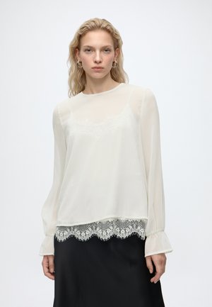 Lyshåret kvinde iført transparent hvid bluse med lange ærmer og blonder i kanten samt sort nederdel, stående mod en ensfarvet lys baggrund.