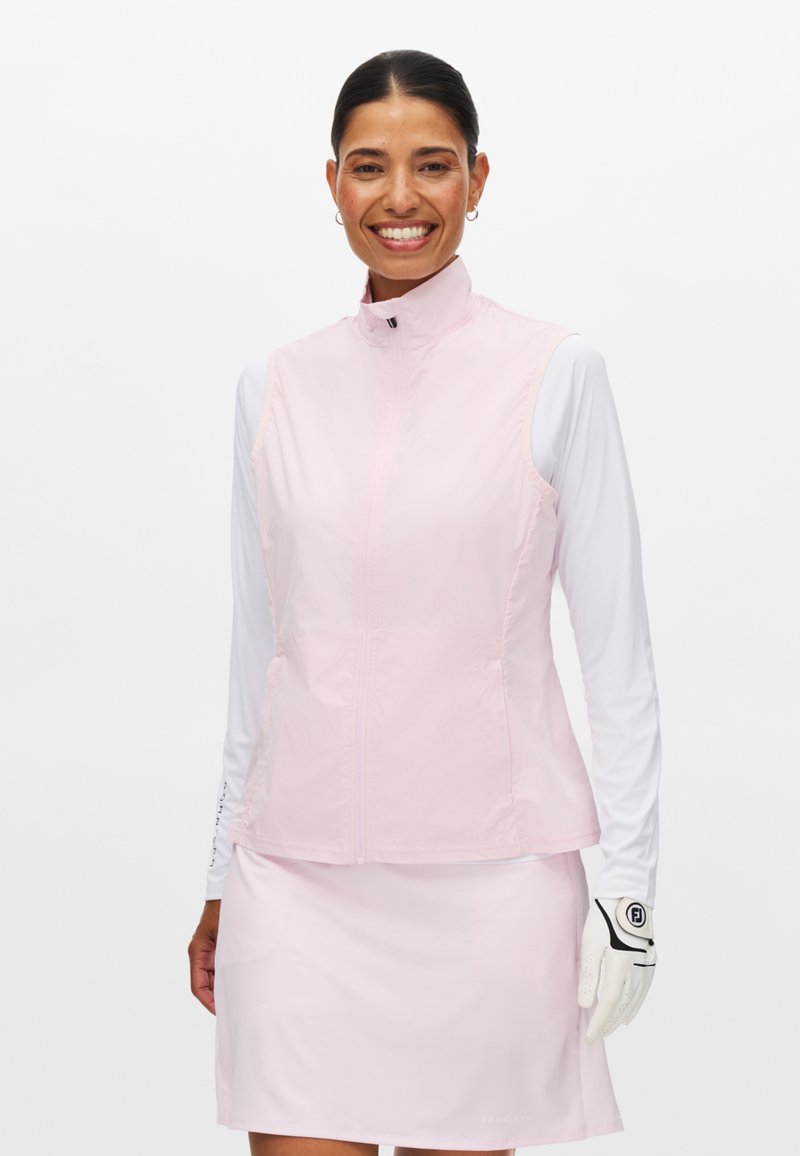 Glunderende vrouw die een lichtroze mouwloze jas draagt, bijpassende rok, wit shirt met lange mouwen en een witte golfhandschoen aan haar rechterhand.